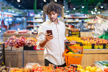 A man looks at his phone while grocery shopping, holding a basket with fresh produce in a supermarket setting.