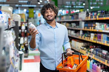A smiling man in a supermarket holds a product while shopping with a basket, surrounded by shelves.