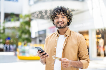 A cheerful man with curly hair smiles while holding a credit card and using his smartphone in a...