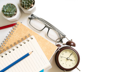 A notebook and pencils sits on a table with an alarm clock and a pair of glasses, isolated on white