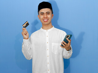 Portrait of a smiling Asian Muslim man holding a credit card and smartphone, ideal for Ramadan and...