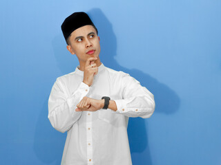 Portrait of a confused young Asian Muslim man wearing traditional clothes, looking at his watch...