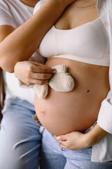 Pregnant woman in blue jeans and white top standing near her partner and holding white baby socks near pregnant belly indoor. Family concep