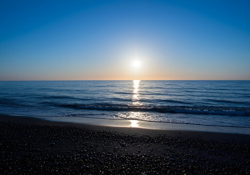 peaceful seascape showing golden sunset over tranquil ocean with sun reflection on water and rocky beach coastline at dusk
