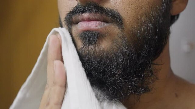 A closeup footage of a young man drying his beard with a white towel, with blurred background. Hygiene concept