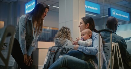 Naklejka premium Crowded International Airport Terminal: Adult Woman Sitting with Infant and Waiting for Airplane Flight. Female Airport Worker Talking to Woman. Diverse Passengers in Departure Lounge of Airline Hub.