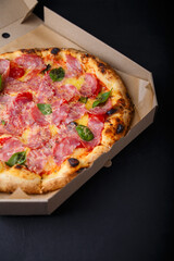 Pizza with cheese, ham, green basil leaves and tomatoes in paper box on black background. Food product advertising banner, copy space