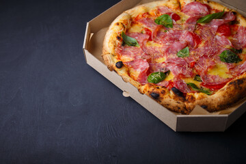 Pizza with cheese, ham, green basil leaves and tomatoes in paper box on black background. Food product advertising banner, copy space