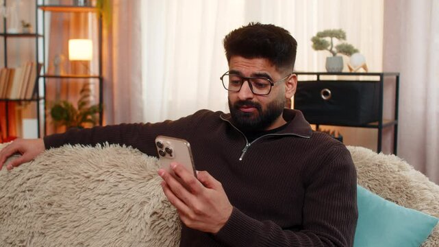 Indian man at home browses ex-girlfriend social media profile with sad eyes and heavy sigh. Arabian guy on sofa puts smartphone aside slowly, feeling nostalgic, disappointed and emotionally tired