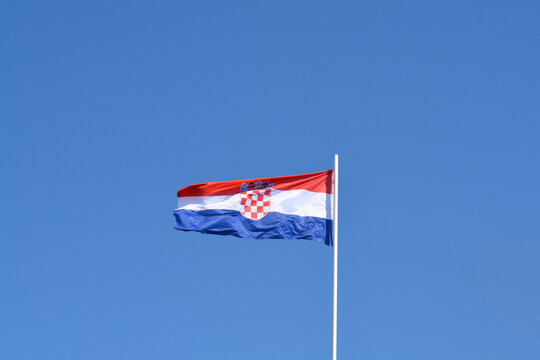 The Croatian national flag, featuring red, white, and blue horizontal stripes and the checkered coat of arms, waves gently against a clear blue sky