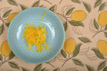 Lemon zest is piled in the center of a light blue patterned plate set on a tablecloth with a lemon...