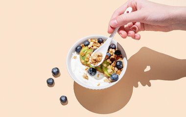 Hand holding spoon natural yogurt with granola, kiwi and blueberry in bowl on beige background