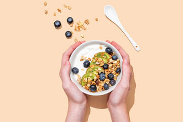 Hands holding bowl of natural yogurt with granola, kiwi and blueberry on beige background.