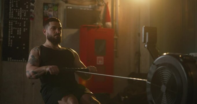 Man with beard and tattoo doing rowing exercises on machine during his strength training in modern cozy fitness room with yellow lighting. Rowing Machine Workout