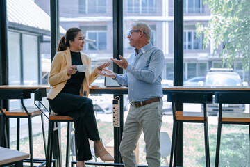 Senior businessman engaging in a discussion with businesswoman, sharing insights while she sips coffee during a break in a modern office cafeteria, fostering collaboration and teamwork