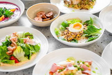 A variety of freshly prepared dishes on a table, including salads, sliced meats, sausages, a soft-boiled egg, and a vibrant soup garnished with herbs and vegetables.