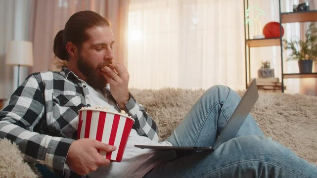Young man eating popcorn at home, occasionally tossing kernels playfully while watching funny clips on laptop. Caucasian guy relaxes comfortably on sofa, laughing out loud at silly and amusing videos