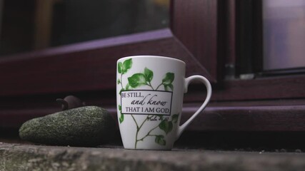 Close-up of hands placing a tea mug on stone step in soft daylight, with words be still and know that I am God