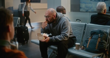 Crowded International Airport Terminal: Man Eating Burger, Waiting for Delayed Airplane Flight,...