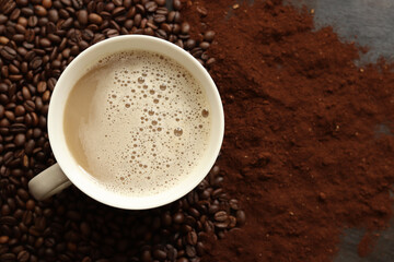 Cappuccino, top view. Cup of cappuccino among coffee beans and ground coffee. Cup of aromatic coffee among roasted coffee beans. Invigorating drink. Copy space
