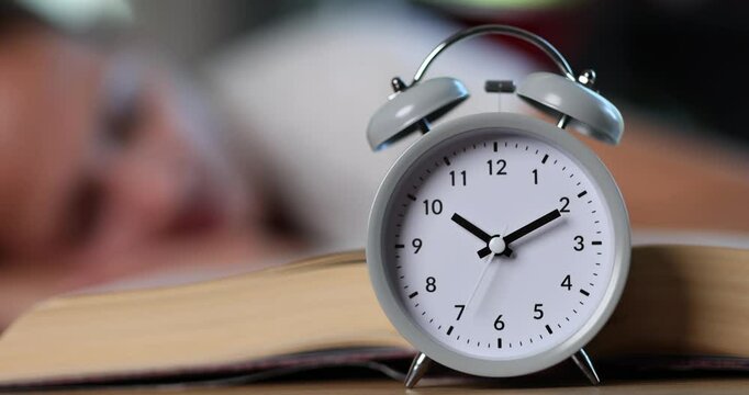 Alarm clock near open book and tired woman student head put on desk. Female overwhelmed by homework and important exam time reminder at home