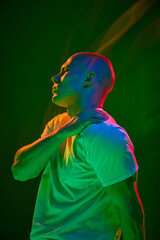 Side view portrait of young bald man in white t-shirt, touching his shoulder and looking to left on...