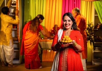 Holding laddu thali with smile, girl invites to join Ganesh puja as family decorates in background