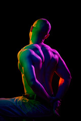 Rear view of young bald man, shirtless, with his hands behind his muscular back, on dark background...