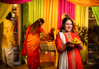 Holding laddu thali with smile, girl invites to join Ganesh puja as family decorates in background