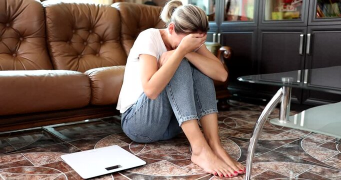 Disappointed woman in casual clothes sits on floor and curls legs to chest crying. Weight scale lies like painful reminder of inner struggle