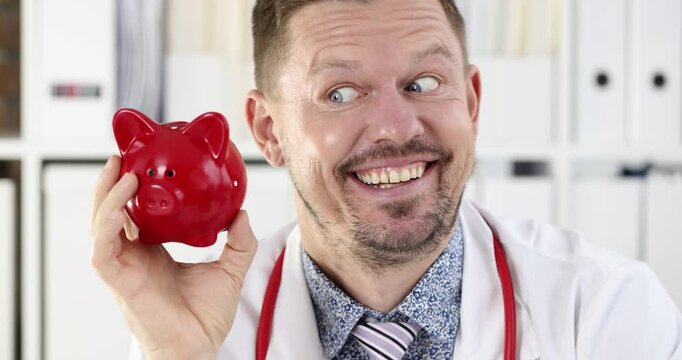 Smiling man doctor shakes red piggy bank symbolizing rising healthcare costs in clinic. Medic reminds about importance of saving for medical expenses