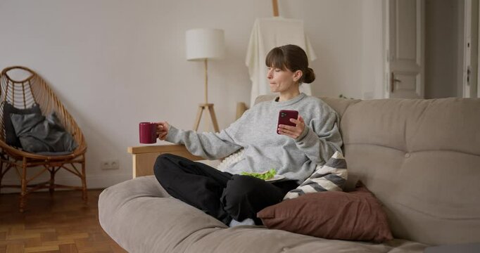 Woman drinking tea and eating sandwich while holding her phone and sitting on the sofa relaxing at home. Rest and leisure with a snack and a phone at home
