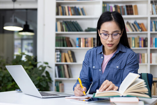 A young woman with glasses studies in a library, using books, a laptop, and taking notes. It captures the essence of learning and research.
