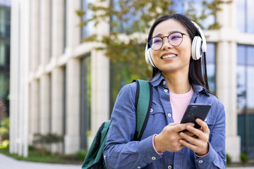 An asian student, wearing glasses and headphones, smiles while looking at her phone outdoors. She...