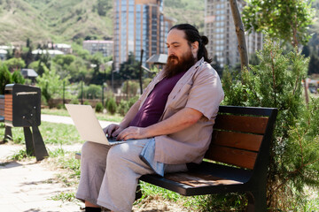 Obraz premium Plus-size man working and relaxing outdoors using a laptop, smartphone, and notebook on a park bench during a sunny day