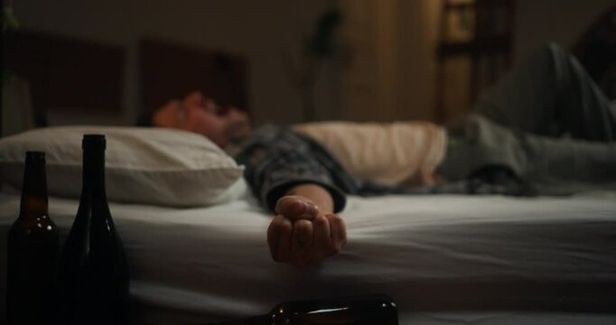 Man in checkered shirt lying on white mattress screaming and trying to rest after despair and drinking alcohol with empty bottles in room. A man on a drinking binge