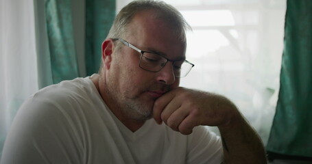Man gazing pensively while resting his chin on his hand, seated near a window with soft green...