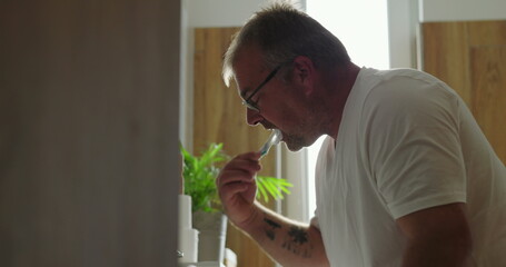 Man brushing teeth at bathroom sink, side profile view with concentrated expression, morning dental...