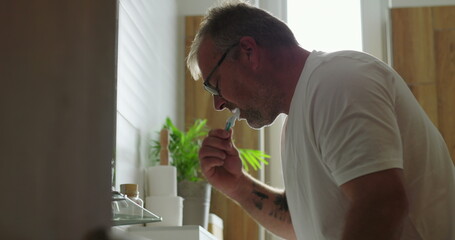 Man brushing teeth at bathroom sink, side profile view with concentrated expression, morning dental...