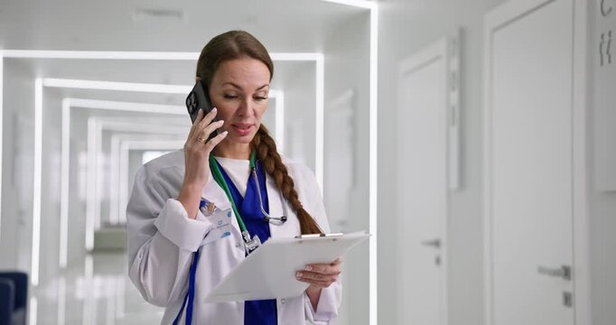 Female doctor in white coat holds tablet with papers and communicates via phone in corridor of modern hospital
