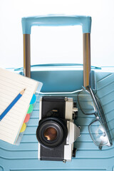 A blue suitcase with a camera, sunglasses and a notebook on top of it. The notebook is on the left side of the suitcase. The camera is a vintage model. white copy space