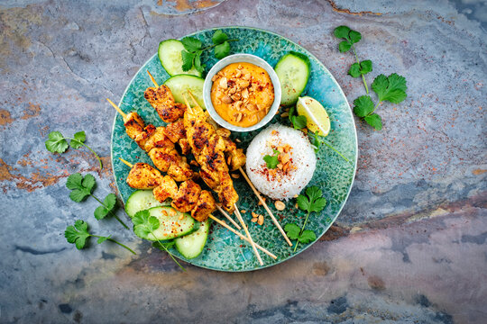 Sate ayam- grilled chicken satay sate skewers with peanut dipping spicy sauce, steamed rice and fresh cucumber served as top view on a design plate