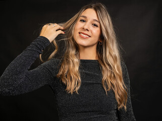 Fototapeta premium Cheerful young woman with long blonde hair playing with her hair while smiling, wearing a sparkly black dress against a dark studio background