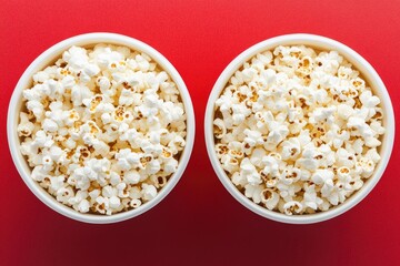 Two white bowls filled with popcorn sit atop a vibrant red surface.  Each bowl is brimming with kernels, showcasing a light golden hue