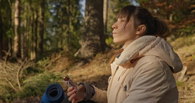 Girl in hiking clothes eats protein bar and drinks tea during her rest stop in sunny spring forest. Snack at a halt During the journey