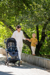 A young mother and her toddler son enjoy a walk in a sunlit park. The child balances on a low stone...