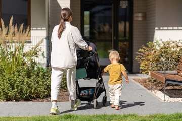 Rear view of a young mother pushing a stroller and her toddler son walking beside her. They are on...