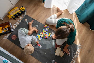 Aerial view of a woman and her young child playing an educational board game with colorful tiles on a space-themed rug. Focus on interactive learning, family bonding, and quality playtime at home.