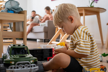 A young child sits on the floor, deeply focused on playing with various toy trucks. Parents relax...