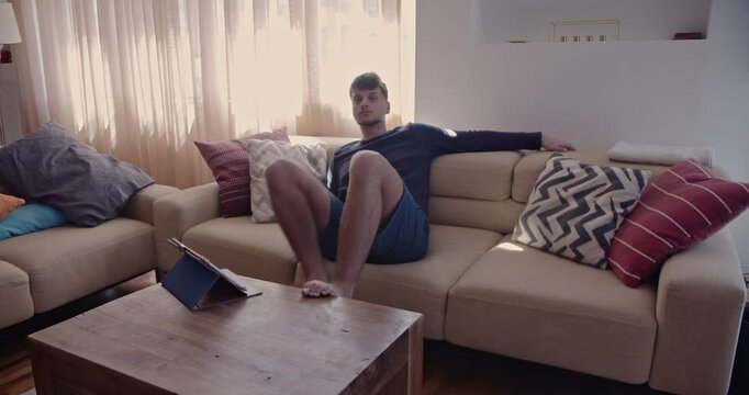 A young man relaxes on his couch, watching television and changing channels with the remote. He puts his feet up on the table and enjoys his leisure time.